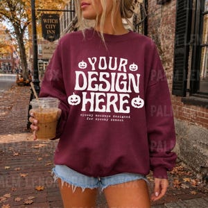 Könnte beinhalten: Ein kastanienbraunes Sweatshirt mit Rundhalsausschnitt, das auf der Vorderseite den weißen Schriftzug YOUR DESIGN HERE, vier kleine Kürbis-Symbole und den kleineren Text spooky mockups designed for spooky season aufweist.