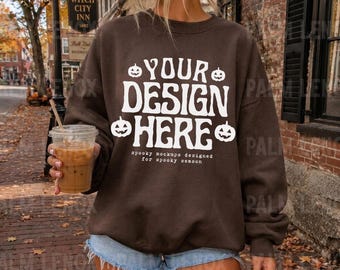 Puede incluir: Una sudadera de cuello redondo color marrón oscuro con texto blanco que dice "YOUR DESIGN HERE" y "spooky mockups designed for spooky season", decorada con cuatro iconos de calabazas blancas alrededor del texto.