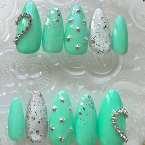 Puede incluir: Un juego de diez uñas postizas con forma de almendra en color verde menta brillante. El diseño presenta detalles de purpurina plateada, pequeños tachones plateados en forma de estrella y una línea curva de pedrería transparente.