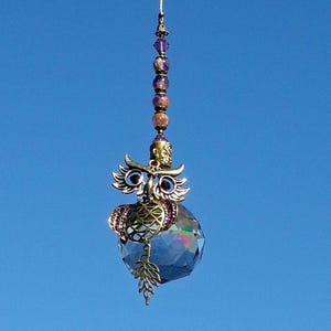 May include: A decorative hanging ornament featuring a bronze-colored owl perched on a faceted crystal ball. The owl has detailed wings and eyes, with a string of beads above. The background is a clear, bright blue sky.