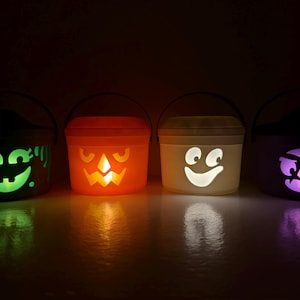 May include: Four plastic Halloween buckets with black handles, each featuring a unique glowing face cutout. The buckets are illuminated from within, displaying green, orange, white, and purple light through the facial expressions of a monster, jack-o'-lantern, ghost, and vampire.