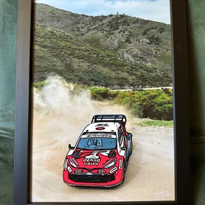 May include: A framed print featuring a red and white rally car kicking up dust on a dirt track. The car displays the text DENSO and TAVADIO on the bonnet. The background shows a green hillside under a blue sky. The artwork is enclosed in a black frame.