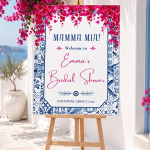 Peut inclure: Un panneau d'accueil rectangulaire sur un chevalet en bois avec un fond blanc, une bordure à motifs de carreaux bleus et blancs et des fleurs de bougainvillier rose vif en haut. Le texte indique Mamma Mia! Welcome to Emma's Bridal Shower Santorini, Greece 2026.