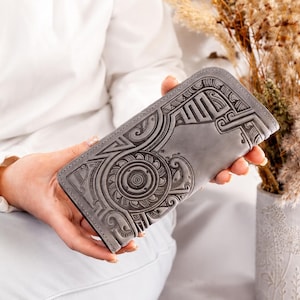 May include: A rectangular grey leather wallet featuring an intricate embossed pattern of circular and geometric motifs. The wallet has a smooth, matte finish and a structured design with rounded corners, showcasing detailed craftsmanship on its surface.