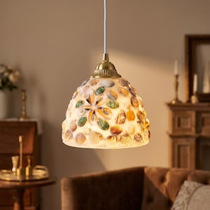 May include: A hanging lampshade decorated with seashells in shades of white, green, and brown. The light fitting has a brass-coloured top and a white cord. The design suggests a coastal or nautical theme, suitable for a living room or bedroom.