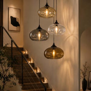 Pode incluir: Quatro luminárias pendentes com abajures ovais, âmbar, transparentes e cinzentos, penduradas no teto. Cada luminária tem uma fixação preta e uma lâmpada Edison visível. As luzes estão suspensas acima de uma escada de madeira.
