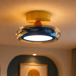 May include: A modern ceiling light fixture with a yellow base, orange accent, and a clear, rounded glass shade with a blue interior. The light emits a bright, white glow, illuminating the room. The fixture is mounted on a white ceiling.