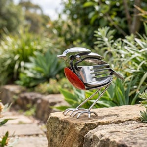 May include: A unique metal bird sculpture made from recycled cutlery. The bird has a polished silver head and body, with a bright red wing. It stands on slender metal legs, positioned on a stone in a garden setting.