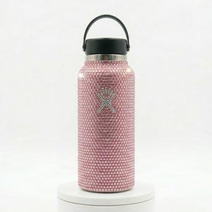 Könnte beinhalten: Eine rosa Wasserflasche, bedeckt mit kleinen, glitzernden Strasssteinen. Die Flasche hat einen schwarzen Deckel und Griff sowie ein silbernes Logo. Die Flasche ist etwa 25 cm hoch.