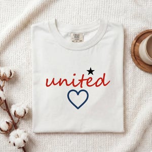 Könnte beinhalten: Weißes T-Shirt mit dem Wort "united" in roter Schrift, einem schwarzen Stern und einem blauen Herzen. Das T-Shirt ist auf einer weißen, strukturierten Oberfläche gefaltet, mit einem Holzuntersetzer und Baumwollstielen im Hintergrund.