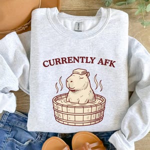 Könnte beinhalten: Ein hellgraues Sweatshirt mit Rundhalsausschnitt und einer Grafik eines Capybaras, das in einem Holzzuber sitzt und ein Handtuch auf dem Kopf trägt. Der Text "CURRENTLY AFK" ist in Dunkelrot über der Illustration gedruckt. Das Sweatshirt hat lange Ärmel.