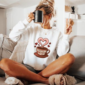 Peut inclure: Sweat-shirt blanc avec un motif de tasse à café avec l'inscription "Brewing Love". Le motif représente une vapeur en forme de cœur et des grains de café. Le sweat-shirt est à manches longues et a un col rond.