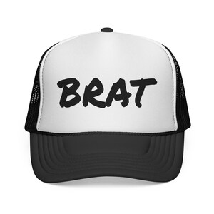 Pode incluir: Um boné trucker preto e branco com um painel frontal branco com a palavra BRAT impressa em letras pretas em negrito. O boné inclui uma aba preta e painéis laterais e traseiros de malha preta, com um botão preto no topo da coroa.