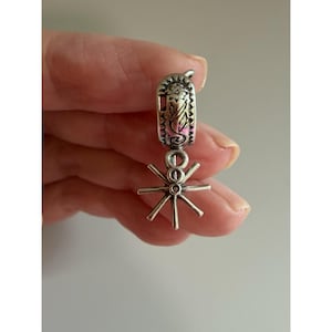 Può includere: Un charm argentato con un design a sperone. Il charm presenta una parte superiore rettangolare con un motivo decorativo e uno sperone pendente con un design a stella. Il charm è progettato per essere attaccato a un braccialetto o una collana.