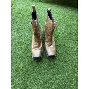 May include: A pair of worn, golden-brown leather ankle boots with square toes. The boots have decorative stitching and pull tabs. The interior is lined with blue fabric. The boots are set on a green, grassy surface.