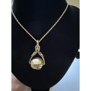 Può includere: Una collana con pendente dorato dal design dettagliato. Il pendente presenta un medaglione ovale con incisioni floreali, sospeso da un'ansa decorativa e una delicata catena. La collana è esposta su una superficie nera.