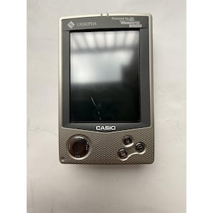 May include: A silver and gray Casio Cassiopeia handheld computer. The device has a screen, a circular control, and several buttons. The screen displays a crack. The text "CASIO" is printed on the device.