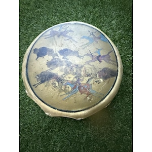 May include: A circular drum featuring a painted scene of bison and riders. The drumhead has a gold-toned background with figures in blue, brown, and red. The drum's frame is a light tan colour. The drum is likely made of animal hide.