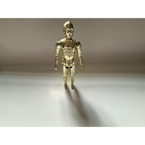 May include: A gold-colored C-3PO action figure from the Star Wars franchise. The figure is standing upright with its arms at its sides. The figure has a detailed design, with visible mechanical components and a smooth, metallic finish. The figure is approximately 6 inches tall.