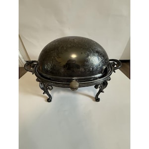 Puede incluir: Un plato de servicio de metal plateado con una tapa abovedada y abatible que presenta intrincados grabados florales. La pieza se apoya sobre cuatro patas ornamentadas y curvas, e incluye dos asas laterales, un pomo frontal y una muesca.