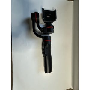 May include: A black handheld gimbal stabiliser with red detailing. The device includes a phone holder and control buttons on the handle. The gimbal is designed to stabilise video recordings, providing smooth footage.