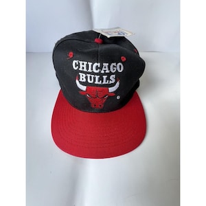 Może przedstawiać: Czarna i czerwona czapka z daszkiem Chicago Bulls. Czapka ma logo drużyny, czerwonego byka i napis "CHICAGO BULLS" w kolorze białym. Daszek jest czerwony, a korona czarna.