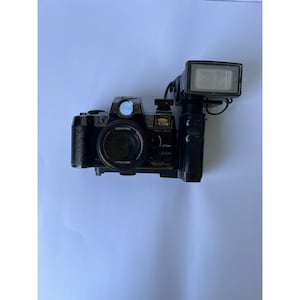 May include: A black Canon QL200 camera with a flash attachment. The camera has a large lens and various dials and buttons. The flash is rectangular and mounted on the top of the camera. The camera is on a white surface.