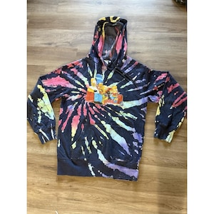 May include: A black tie-dye hoodie featuring a colorful design with pink, yellow, and blue hues. The front of the hoodie displays a graphic of the Simpson family. The hoodie has a drawstring hood and a front pocket.