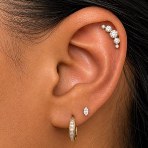 May include: A gold ear cartilage piercing featuring a curved row of five graduated round clear stones. Below, a gold hoop earring with small clear stones and a single marquise-cut clear stone stud earring are worn in the earlobe piercings.