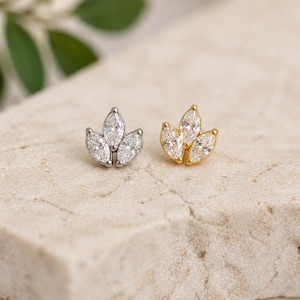 May include: Two stud earrings, one silver-toned and one gold-toned, each featuring a cluster of three marquise-cut clear gemstones. The earrings are displayed on a textured, light-colored surface, with a blurred green leaf in the background.