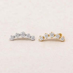 May include: Two curved ear climbers featuring five round-cut clear stones set in metal. One piece is finished in a bright silver tone, while the other is finished in a warm gold tone. Each piece follows a graduated arc design with a central stone.