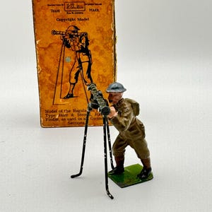 May include: A painted metal toy soldier figure in a brown uniform and blue helmet, operating a black tripod-mounted rangefinder. Behind the figure stands an orange cardboard box with black text and an illustration of the same soldier and equipment.