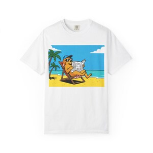 Peut inclure: T-shirt blanc avec un dessin animé d'un chien lisant un journal sur une plage. Le chien porte des lunettes de soleil et une casquette bleue, assis dans une chaise longue. Le fond montre une scène de plage avec des palmiers, du sable, la mer et le ciel.