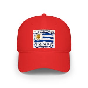 Pode incluir: Boné de beisebol vermelho brilhante com um desenho da bandeira uruguaia, o texto "FIFA WORLD CUP 2026" e "URUGUAY". O design inclui um sol e listras azuis.