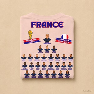 以下が含まれることがあります： 青と赤の「FRANCE」の文字、2026年ワールドカップのグラフィック、円形のフランス国旗がプリントされたライトピンクのTシャツ。その下には、ディディエ・デシャン監督とフランス代表サッカーチームの選手たちのポートレートと名前が青色で印刷されています。