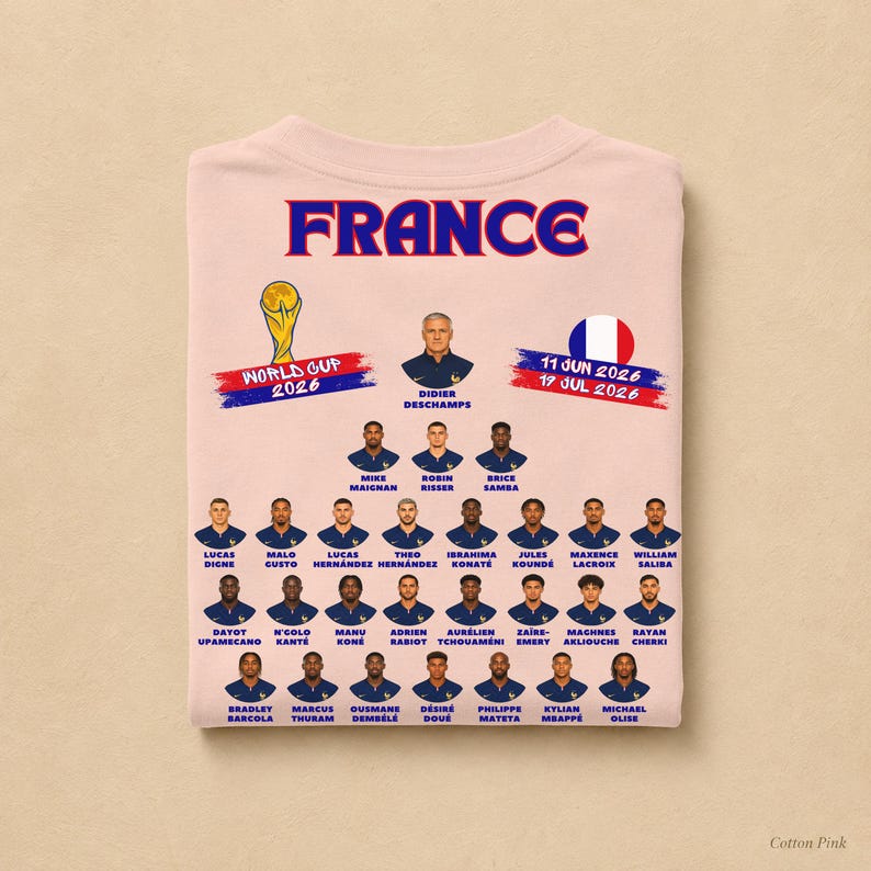 以下が含まれることがあります： 青と赤の「FRANCE」の文字、2026年ワールドカップのグラフィック、円形のフランス国旗がプリントされたライトピンクのTシャツ。その下には、ディディエ・デシャン監督とフランス代表サッカーチームの選手たちのポートレートと名前が青色で印刷されています。