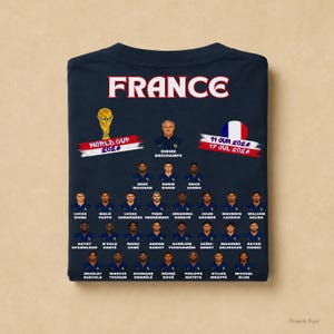 以下が含まれることがあります： 上部に「FRANCE」の文字、ワールドカップ2026のグラフィック、円形のフランス国旗がプリントされたダークネイビーのTシャツ。その下には、フランス代表サッカーチームの選手とディディエ・デシャン監督のポートレートと名前が記載されています。