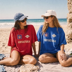 Two women sitting on a sunny beach wearing oversized Hrvatska graphic T-shirts in red and royal blue. Each shirt features a small Croatian-style crest badge, a vintage Hrvatska patch, and bold front lettering in a clean football supporter layout. This lifestyle photo shows the relaxed summer fit, beach styling, casual fan outfit idea, and how the Croatia-inspired tee works for travel, match days, vacations, and everyday streetwear.