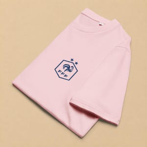 以下が含まれることがあります： 胸元にダークブルーのグラフィックが施されたライトピンクの半袖Tシャツ。グラフィックは、雄鶏のシルエット、FFFの文字、そして六角形の上部頂点の上に配置された2つの小さな星を含む六角形で構成されています。