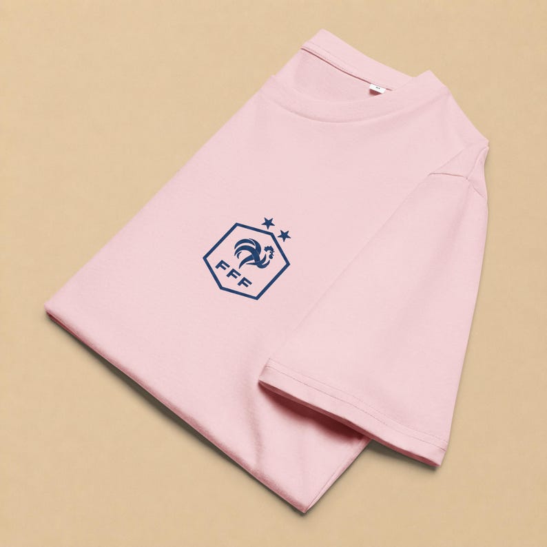 以下が含まれることがあります： 胸元にダークブルーのグラフィックが施されたライトピンクの半袖Tシャツ。グラフィックは、雄鶏のシルエット、FFFの文字、そして六角形の上部頂点の上に配置された2つの小さな星を含む六角形で構成されています。
