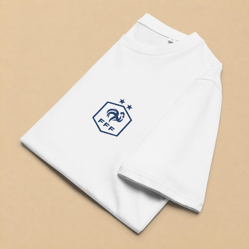 以下が含まれることがあります： 胸元にダークブルーのグラフィックがプリントされた白い半袖Tシャツ。グラフィックは、雄鶏のアイコン、FFFの文字、そして六角形の上に配置された2つの小さな星で構成されており、すべてダークブルーのインクで描かれています。