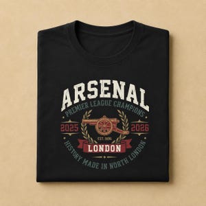 May include: A black t-shirt featuring a graphic design with the text ARSENAL PREMIER LEAGUE CHAMPIONS 2025 2026 LONDON EST. 1886 HISTORY MADE IN NORTH LONDON. The design includes a central cannon illustration surrounded by laurel leaves in red and green.