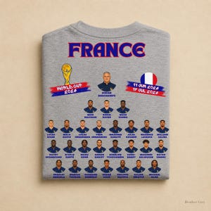 以下が含まれることがあります： 2026年ワールドカップに向けたフランス代表サッカーチームのメンバーがプリントされたヘザーグレーのTシャツ。デザインには「FRANCE」の文字、ワールドカップのトロフィー、フランス国旗、選手やスタッフのポートレートが含まれています。