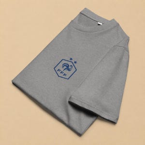 以下が含まれることがあります： 胸元にダークブルーのグラフィックが施された、ヘザーグレーの半袖Tシャツ。グラフィックは、雄鶏のシルエット、FFFの文字、そして六角形の上端の上に配置された2つの小さな星を含む六角形で構成されています。