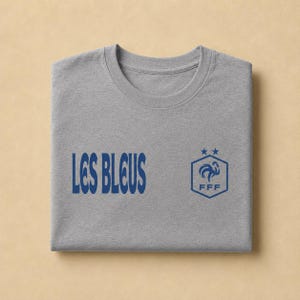 以下が含まれることがあります： ヘザーグレーのTシャツで、左側にダークブルーの「LES BLEUS」という文字、右側にダークブルーの六角形のロゴがプリントされています。ロゴには様式化された雄鶏、FFFの文字、そして六角形の上に配置された2つの小さな星が描かれています。
