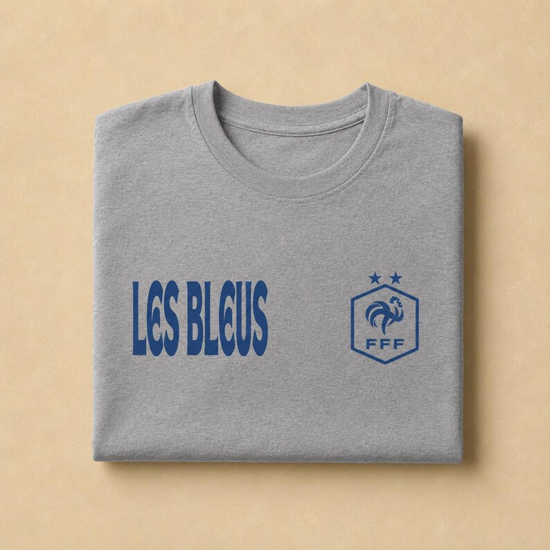 以下が含まれることがあります： ヘザーグレーのTシャツで、左側にダークブルーの「LES BLEUS」という文字、右側にダークブルーの六角形のロゴがプリントされています。ロゴには様式化された雄鶏、FFFの文字、そして六角形の上に配置された2つの小さな星が描かれています。
