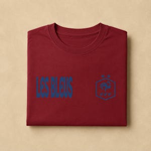 以下が含まれることがあります： クルーネックのマルーンカラーのTシャツ。左側に青い文字で「LES BLEUS」、右側に青い六角形のロゴがプリントされています。ロゴには、様式化された雄鶏のグラフィック、文字「FFF」、六角形の上に配置された2つの小さな星が含まれています。