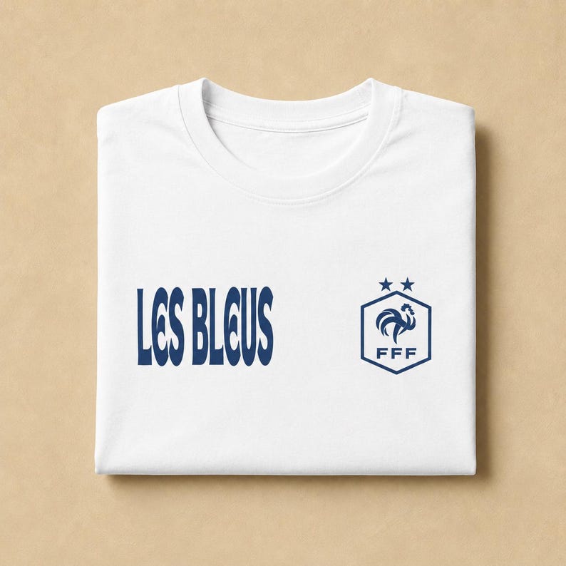 以下が含まれることがあります： 左側に濃い青色の「LES BLEUS」という文字、右側に濃い青色の六角形のロゴがプリントされた白い半袖Tシャツ。ロゴは六角形の中に雄鶏が描かれており、その下に「FFF」の文字、上に2つの星が配置されている。