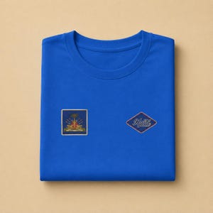 May include: A royal blue crew neck t-shirt featuring two embroidered patches on the front. The left patch is a square displaying the coat of arms of Haiti, while the right patch is a diamond shape containing the word Haiti in a cursive script.