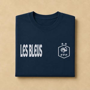 以下が含まれることがあります： 左側に白い文字で「LES BLEUS」、右側に白い六角形のロゴがプリントされたネイビーブルーのTシャツ。ロゴには、様式化された雄鶏、文字「FFF」、そして六角形の形状の上に配置された2つの星が描かれています。